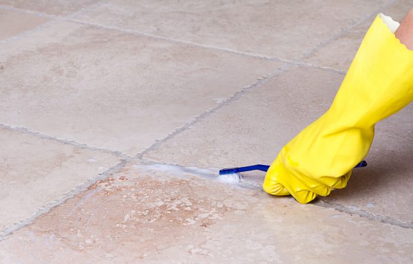 gloved hand cleaning tile floor grout with hydrogen peroxide and a toothbrush