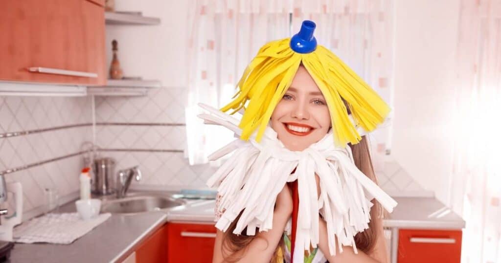 woman being silly wearing mop heads for hair and a beard while trying to get motivated to clean her kitchen